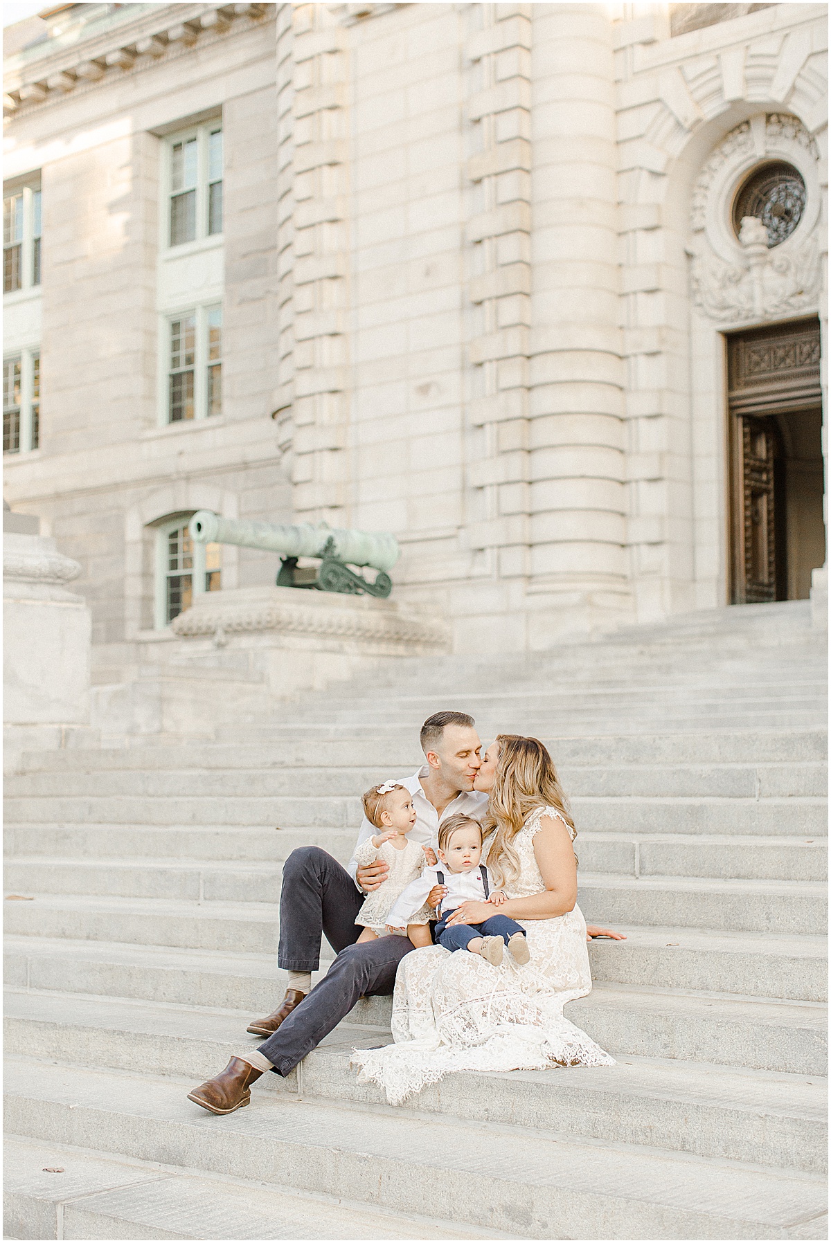 Mom and Dad kissing while holding their children on the steps of a U.S. Naval Academy building in downtown Annapolis Maryland
