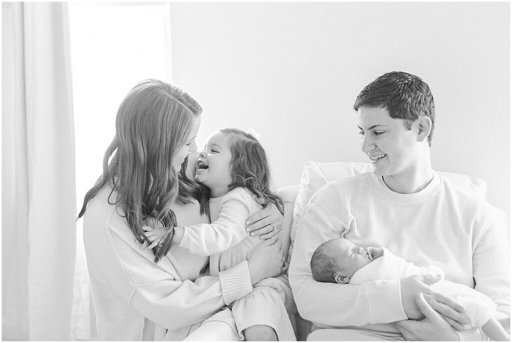 In-home newborn photography image of dad holding baby, and big sister laughing at mom